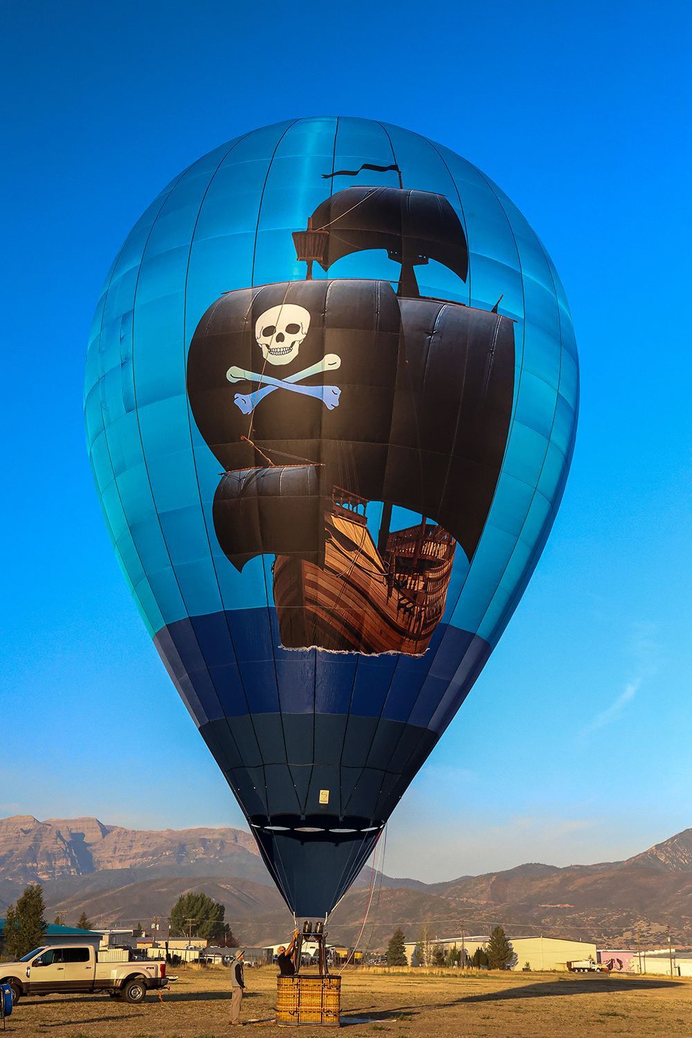 Pirate ship hot air balloon, blue and black, rises against a clear sky over a field.