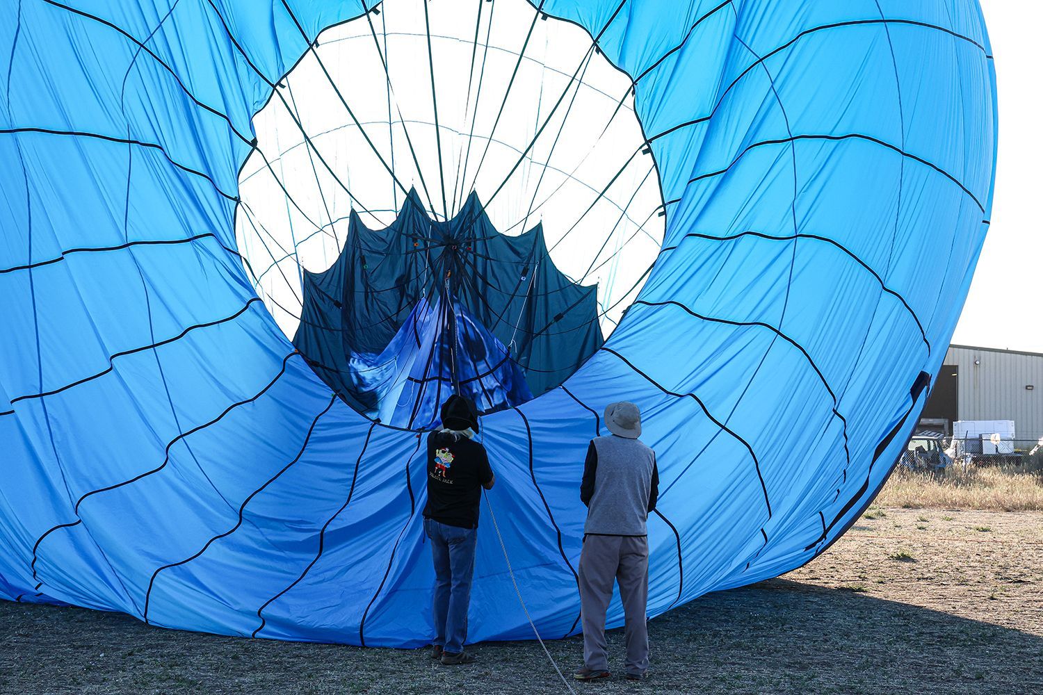 Two people stand inside a blue hot air balloon, watching the flame fill it.