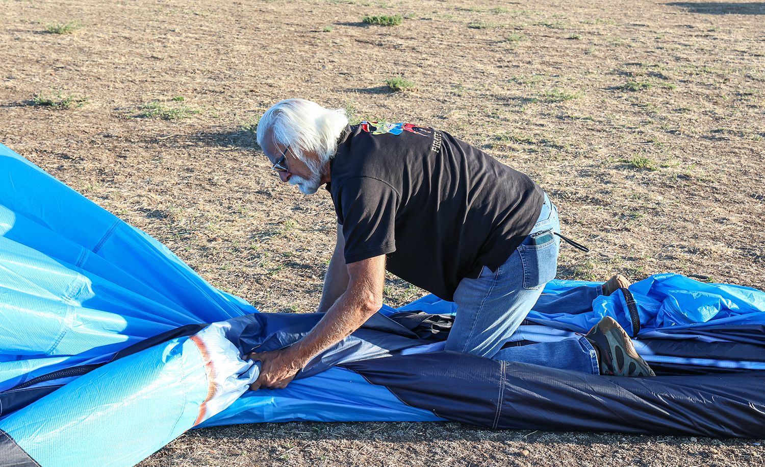 An older man with white hair kneels outdoors, assembling a blue and black tent.