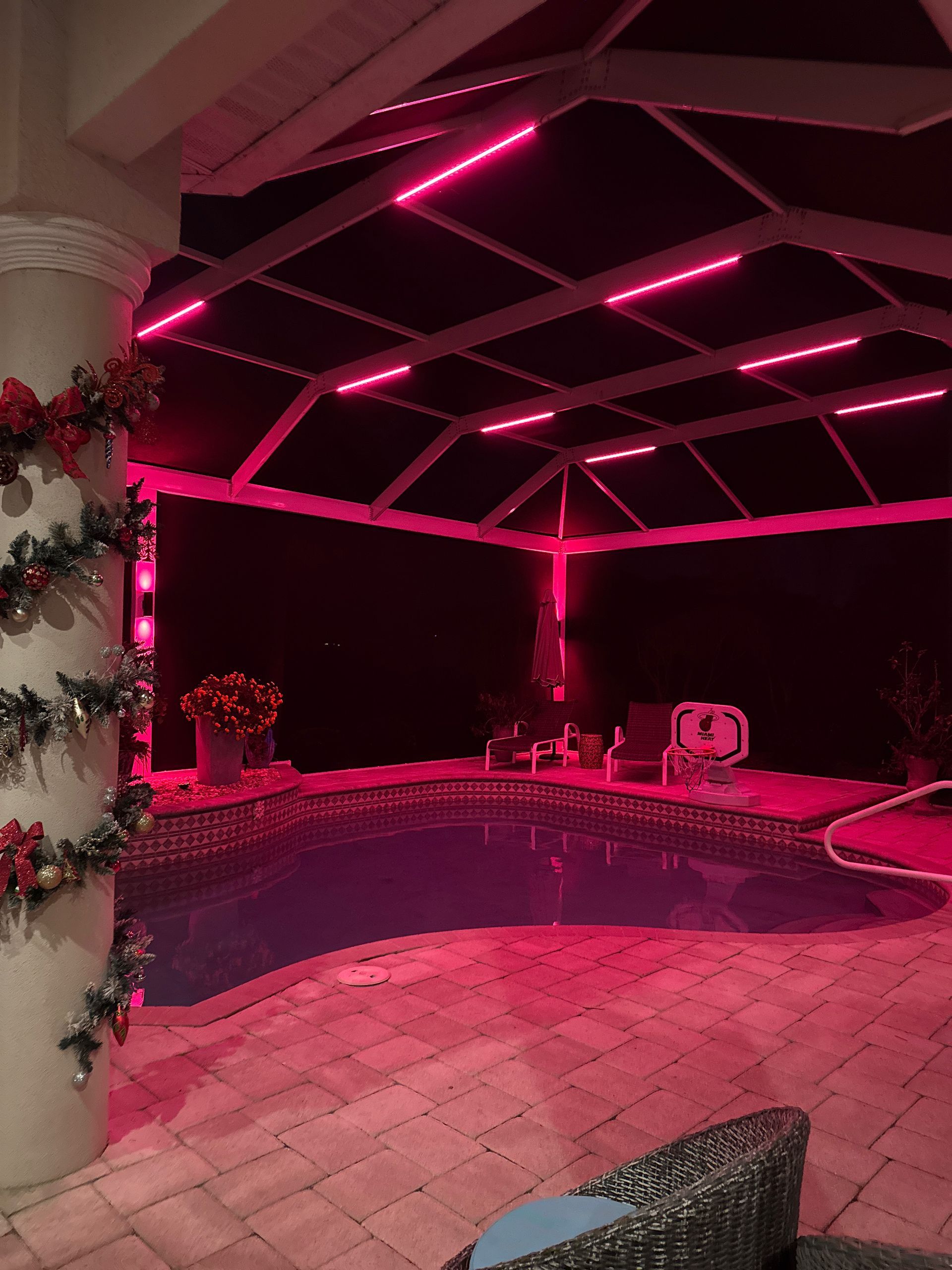 A pool at night with pink LED lights outlining a screened enclosure, a column with festive decor.