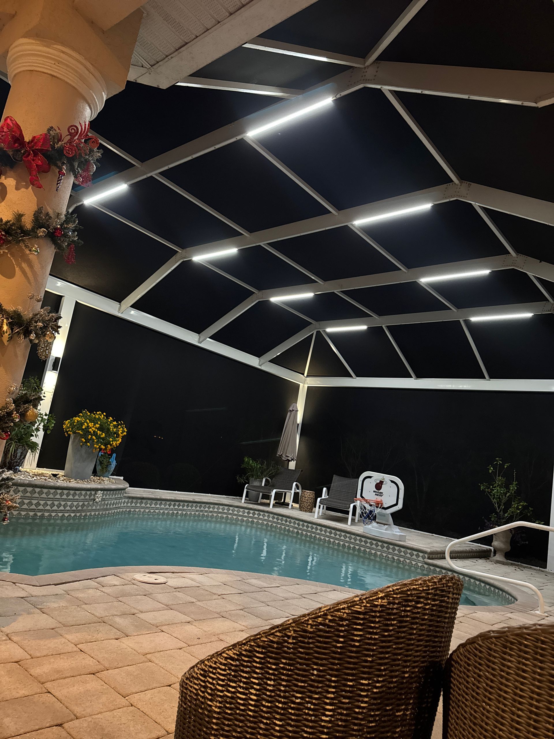 Night view of a pool under a screened enclosure, lit by overhead lights. Wicker chairs in foreground.