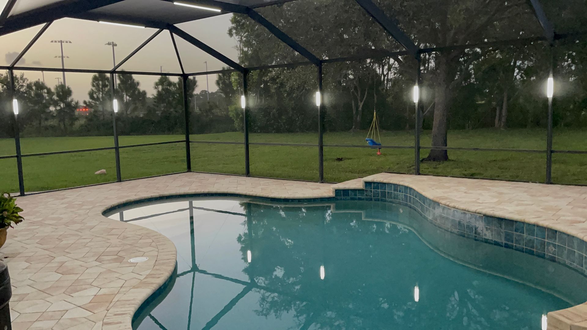 Backyard pool with stone deck, screened enclosure, and grassy lawn at dusk