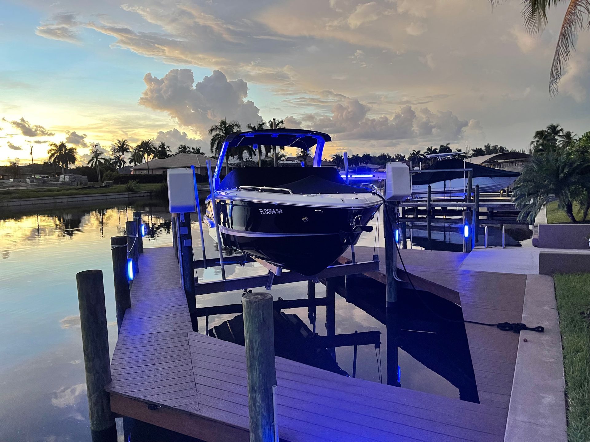 Boat on a lift at sunset, illuminated by blue lights. Canal setting, with palm trees and houses.