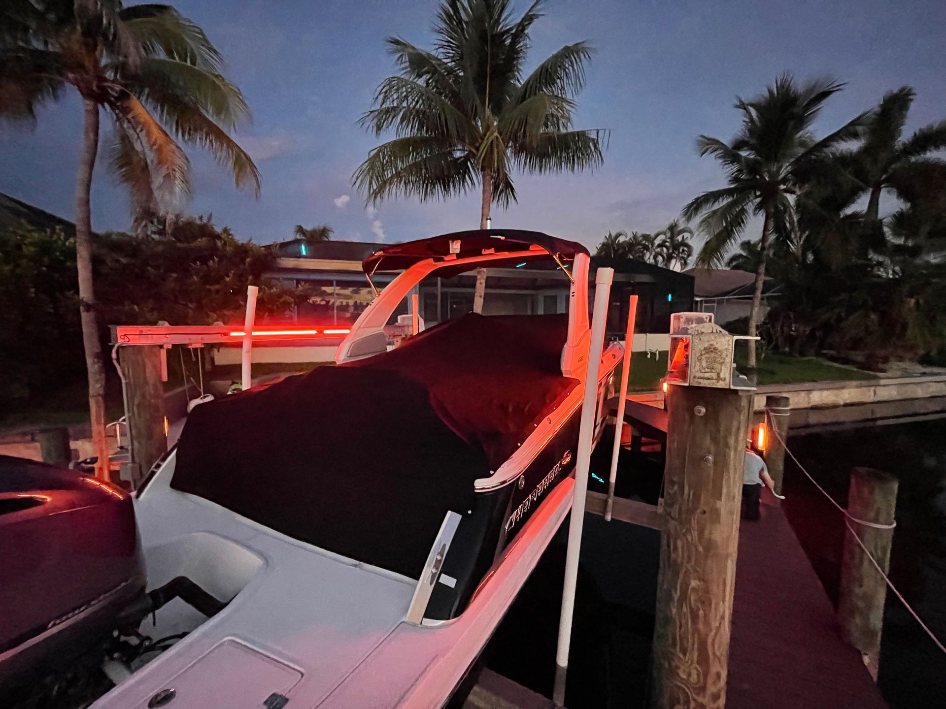 Boat covered with black tarp on a lift near a dock at dusk. Lit with red lights, palm trees in the background.