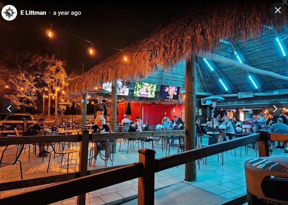 Outdoor bar at night with thatched roof, lights, tables, and people socializing.