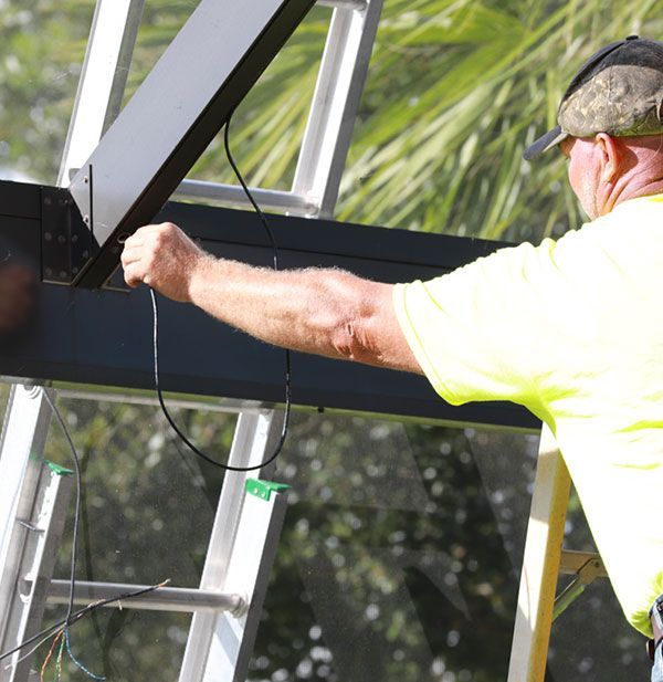 Man in a cap and neon shirt on a ladder, wiring a black metal structure with clear panels.