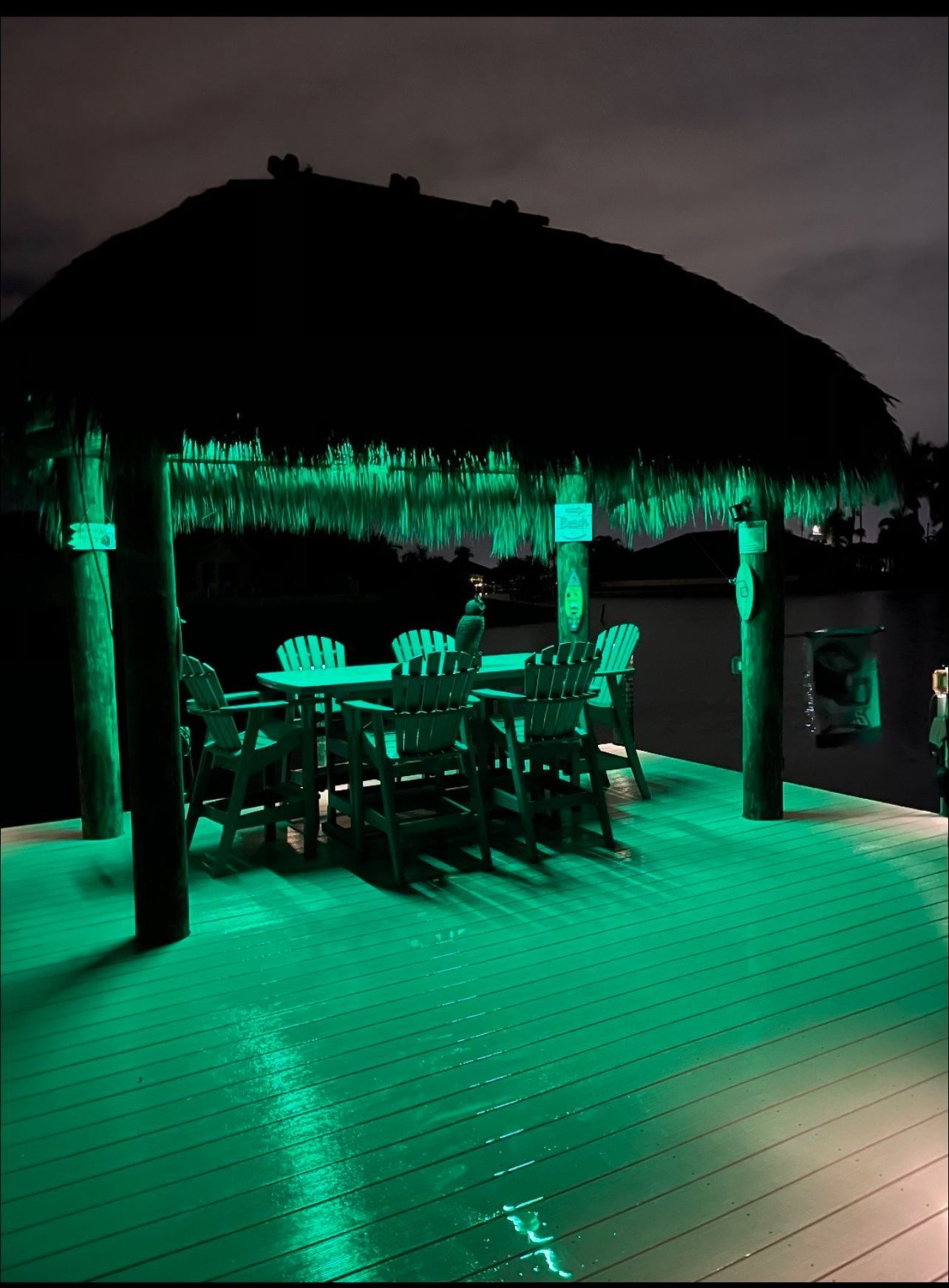 A tiki hut with green lights illuminates a deck and outdoor dining set at night.