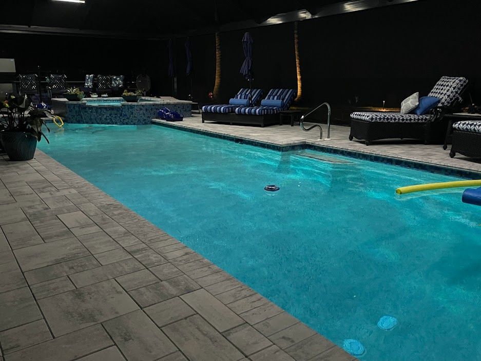 Indoor swimming pool with lounge chairs and hot tub. Bright blue water, gray stone deck, dark walls.
