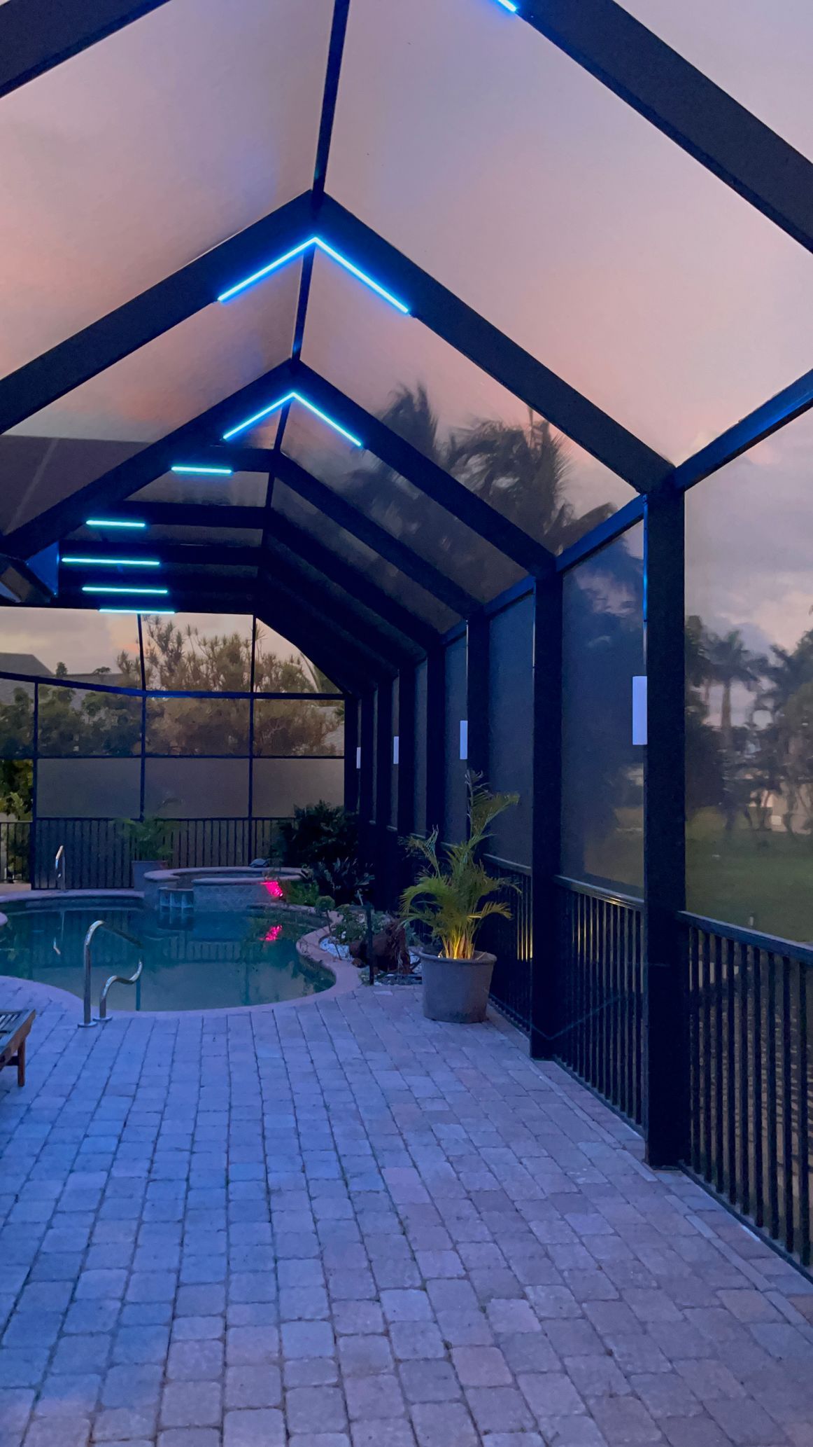 Covered patio walkway beside a pool, with blue-lit beams and a metal railing at dusk
