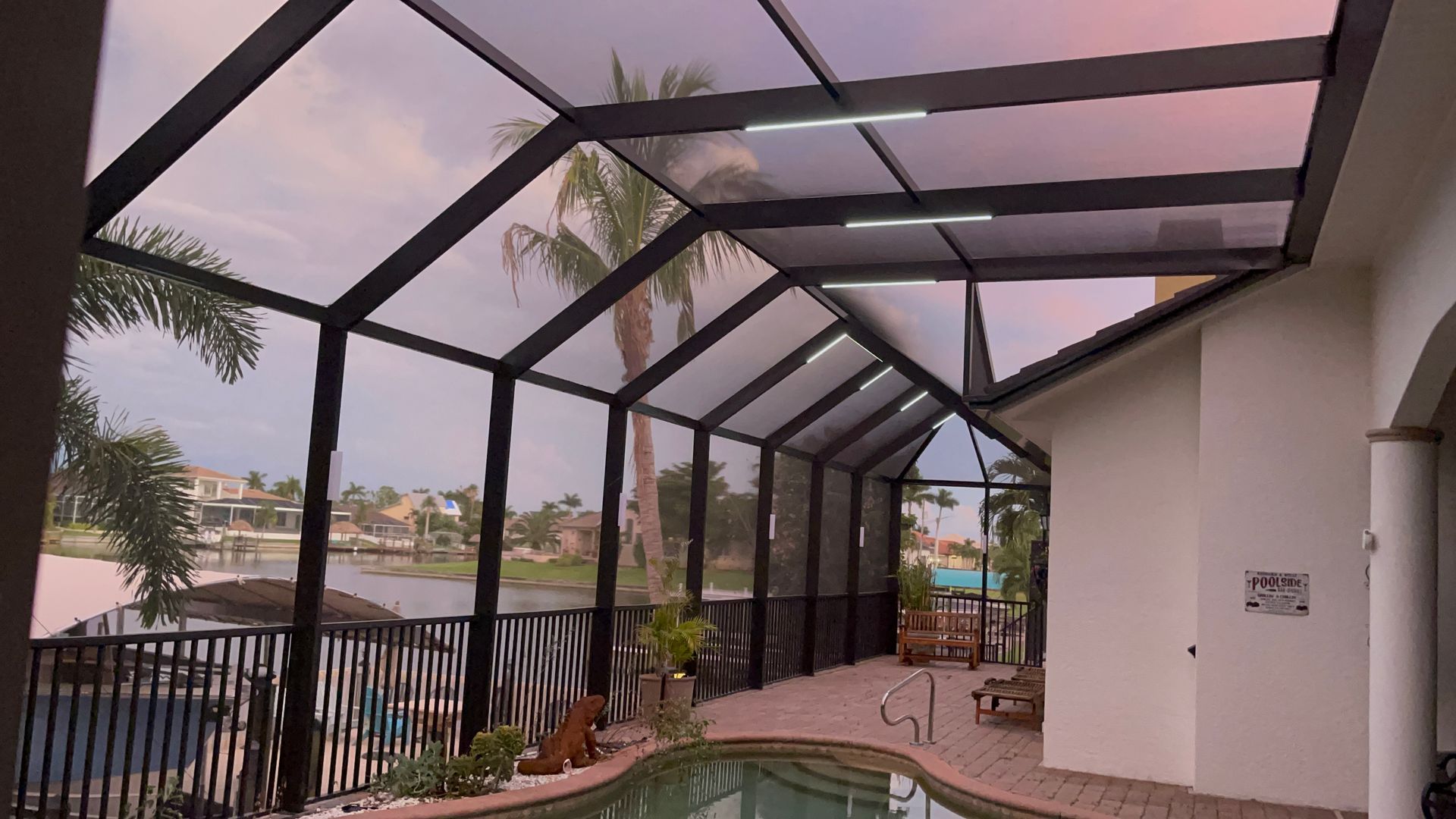 Screened-in pool enclosure with black framing, clear panels, and a view of a waterway and houses at sunset.