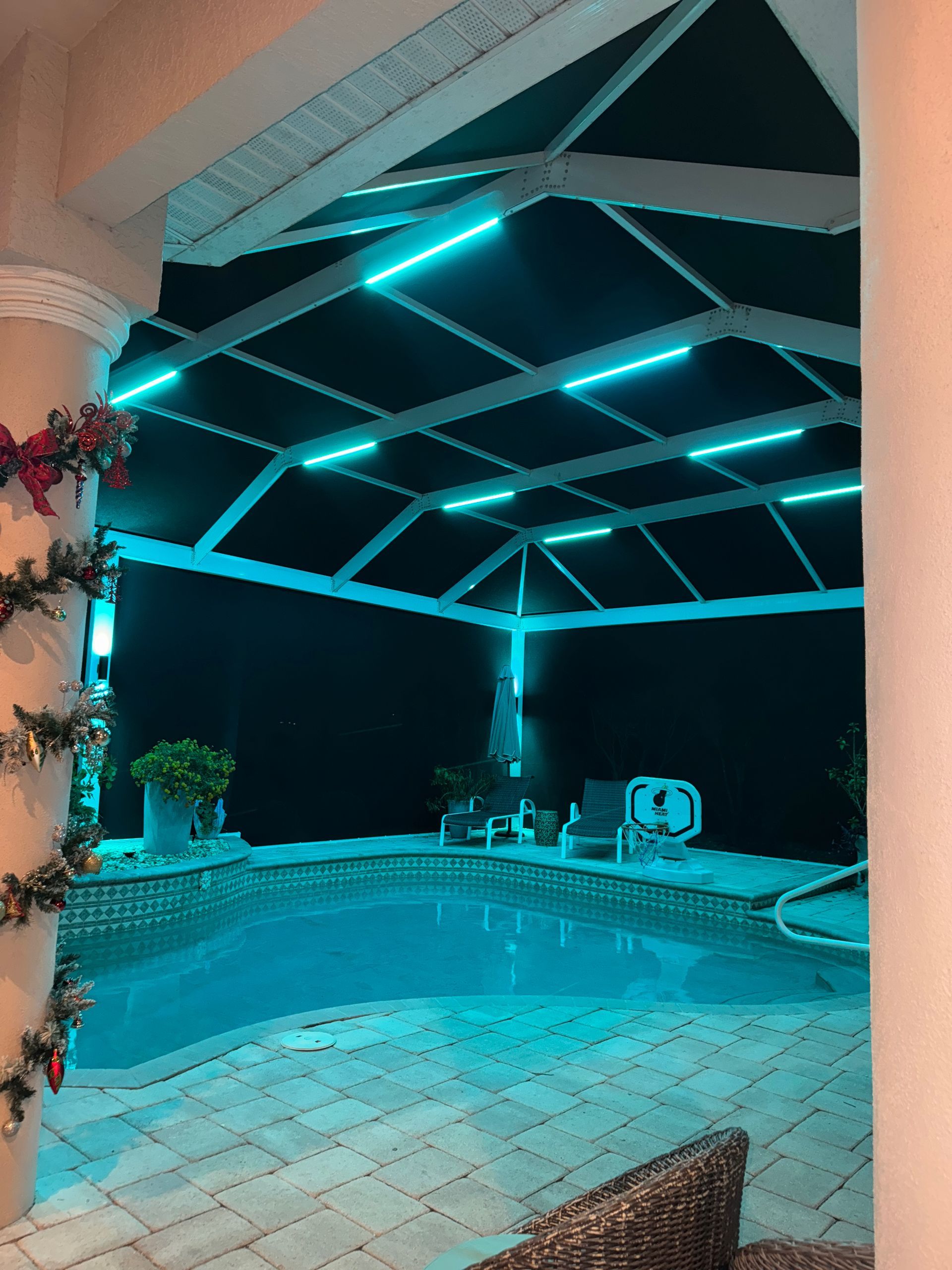 Pool area at night with teal lights on the enclosure and a Christmas garland on a pillar.