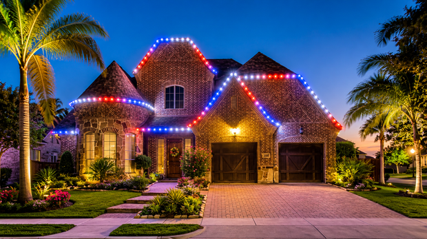 Large decorated house at dusk with colorful roof lights, palm trees, and a lit driveway
