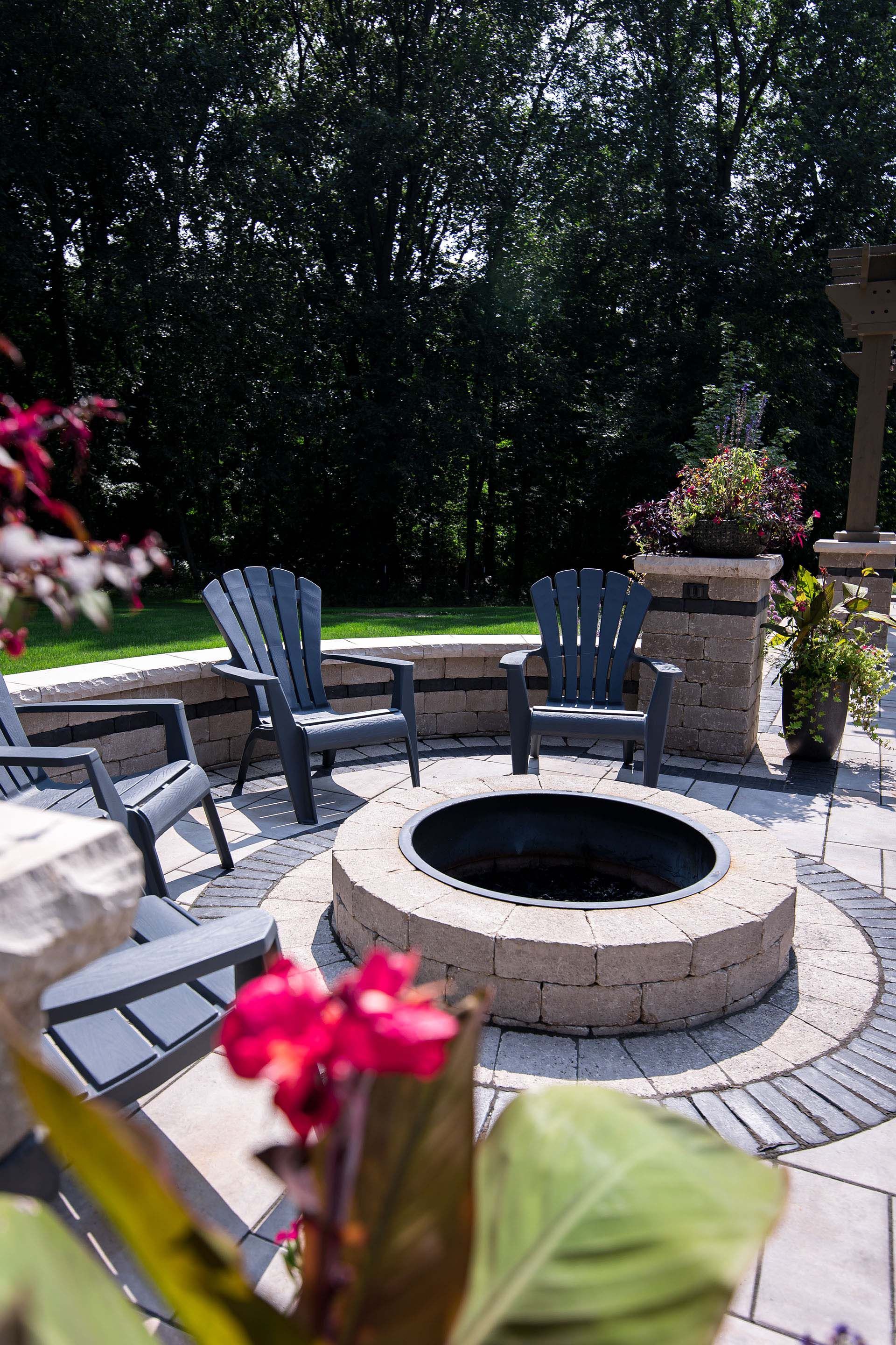 A fire pit is surrounded by chairs and flowers on a patio.