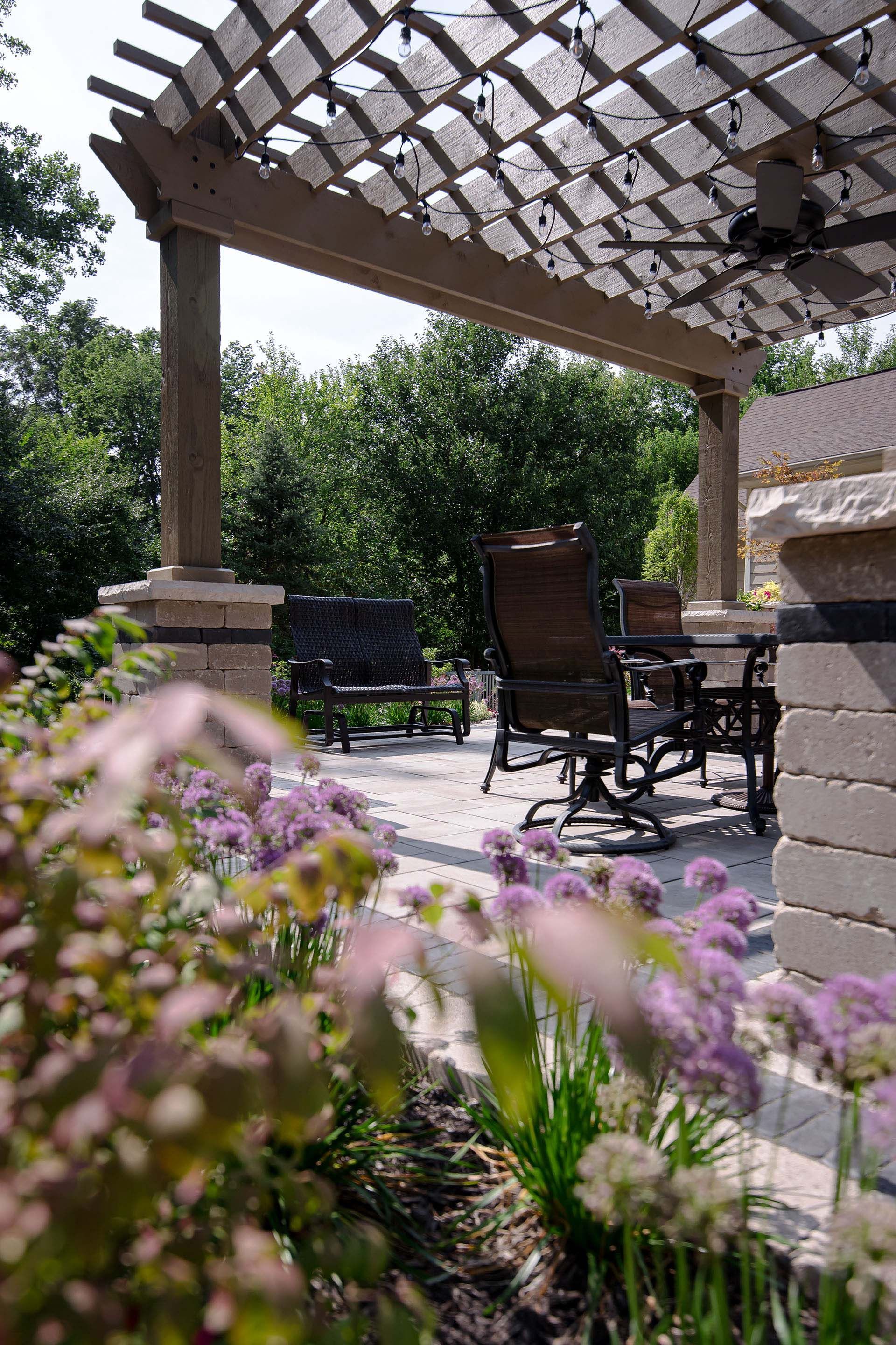 A patio with a pergola and chairs and flowers in the foreground.