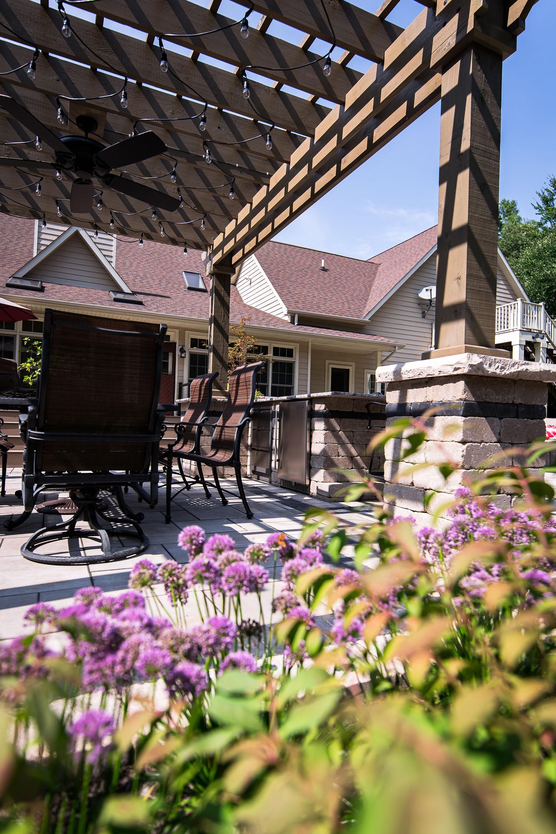 A patio with a table and chairs under a pergola with purple flowers in the foreground.