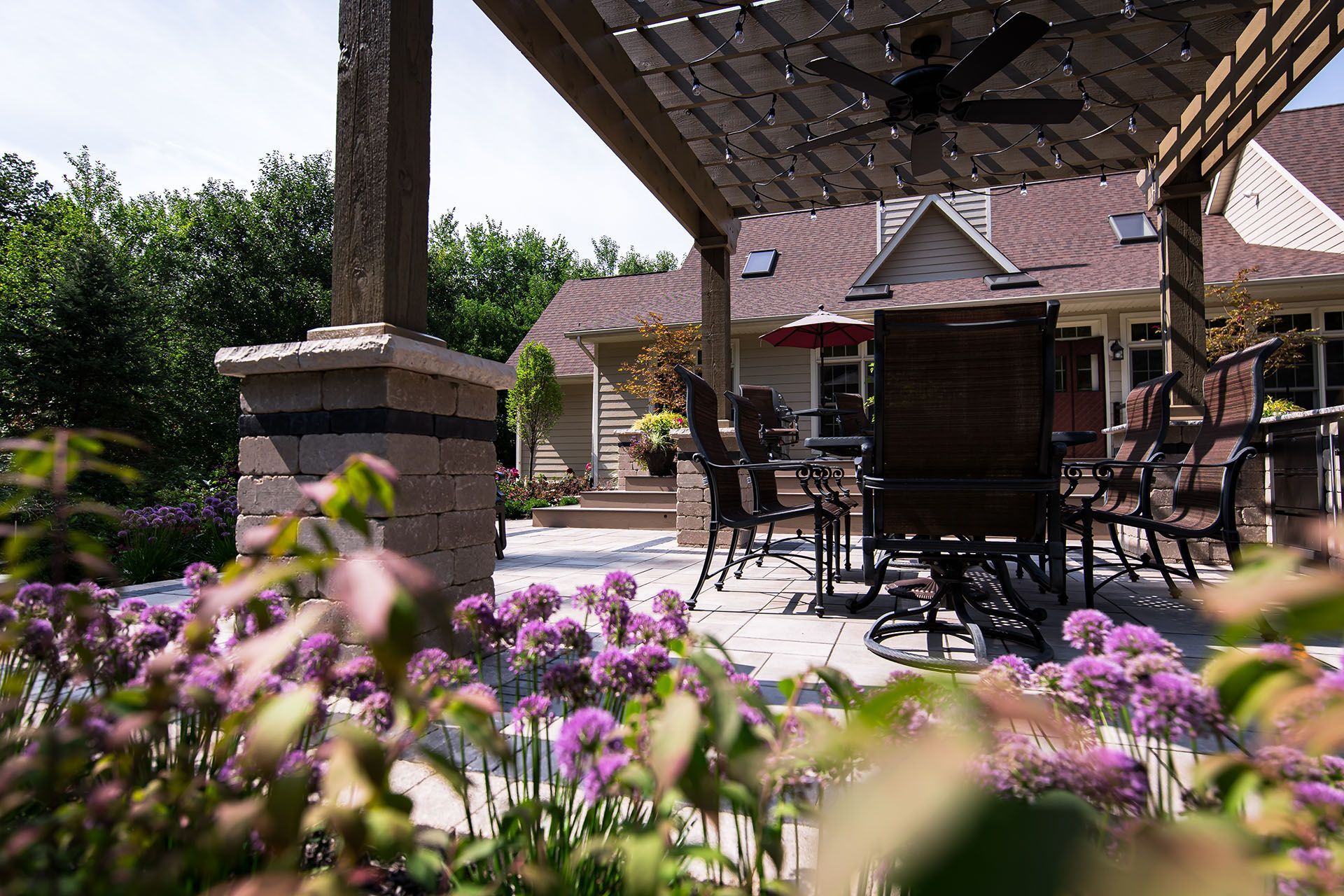 A patio with a table and chairs under a pergola with purple flowers in the foreground.