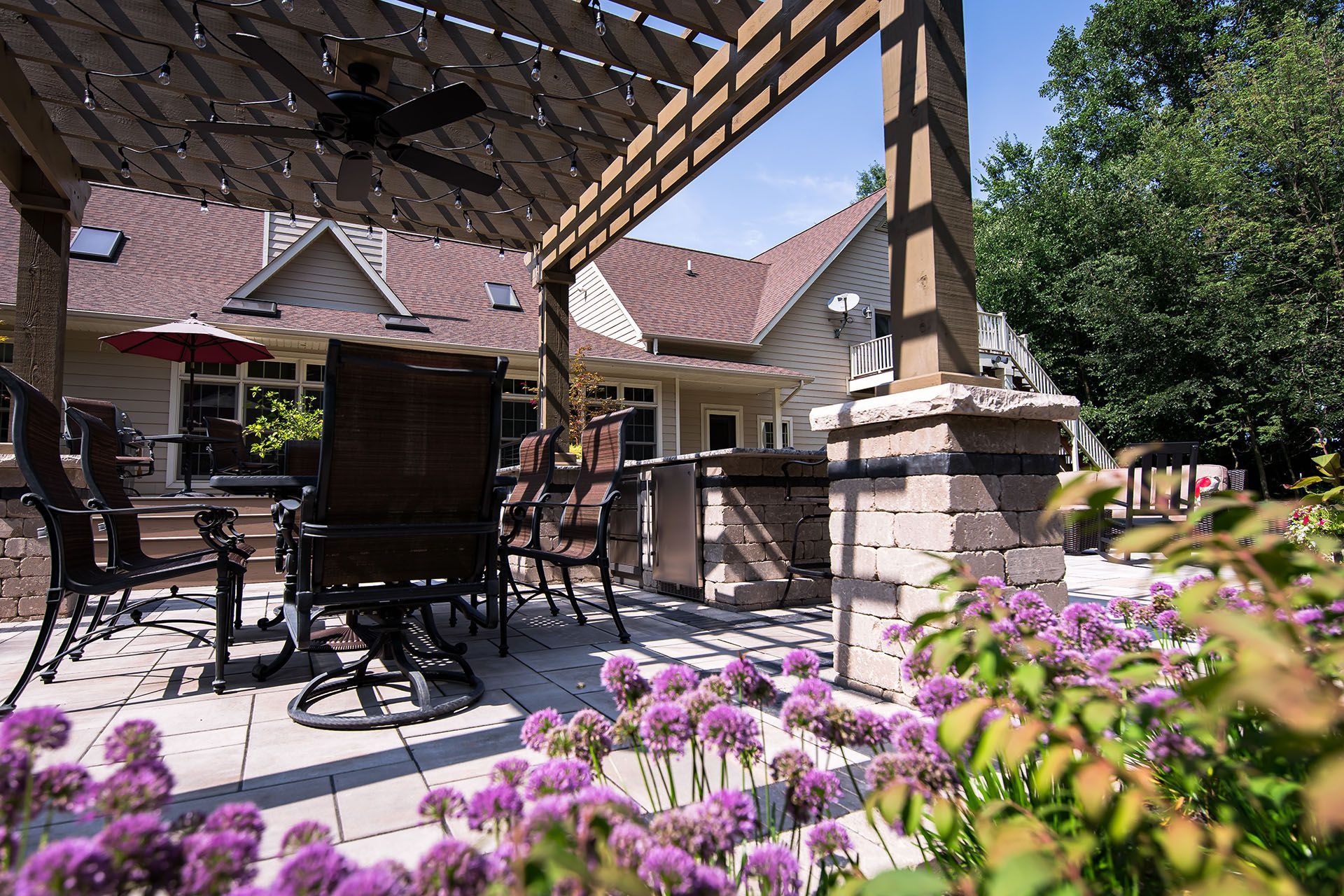 A patio with a table and chairs under a pergola with purple flowers in the foreground.