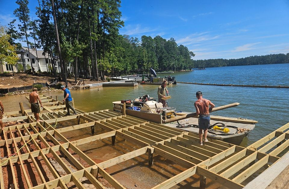 Workers building a wooden dock on a lake. Bright blue sky, green trees, and water surround the structure.