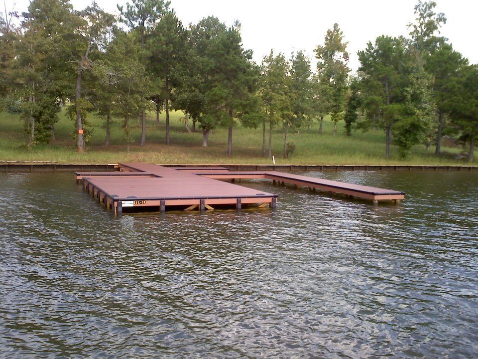 Brown dock on water with trees and grassy bank in the background.