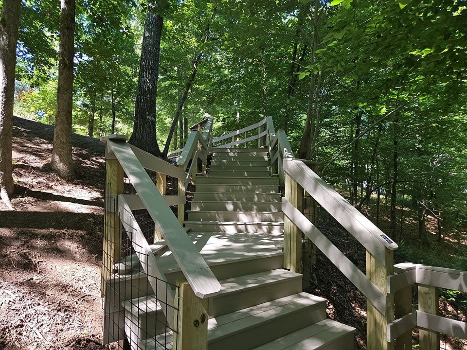 Wooden staircase ascends through a wooded area. Railings with mesh fencing on sides.