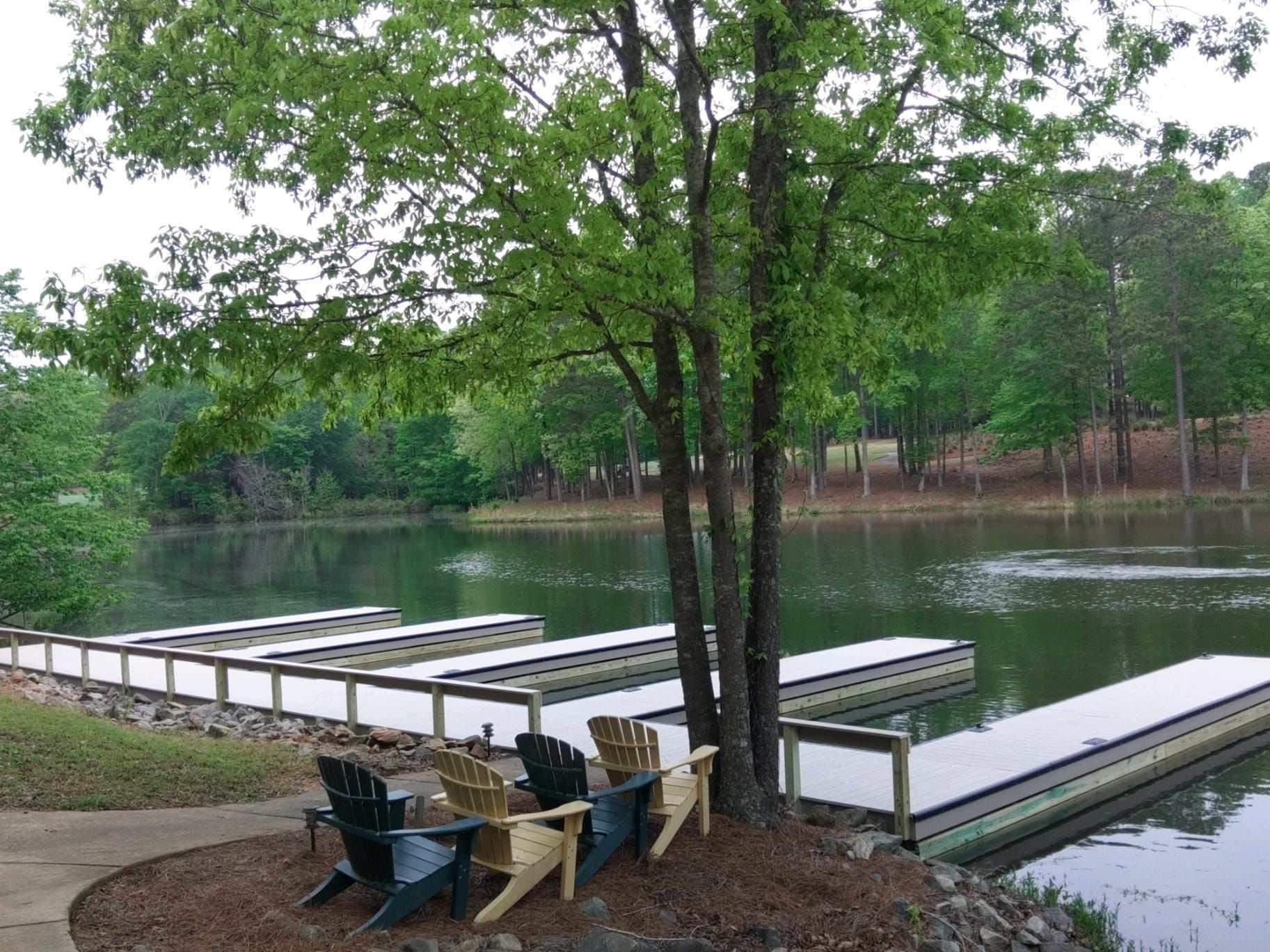 Lakeside dock with several chairs under a tree. Water, trees, and cloudy sky in background.