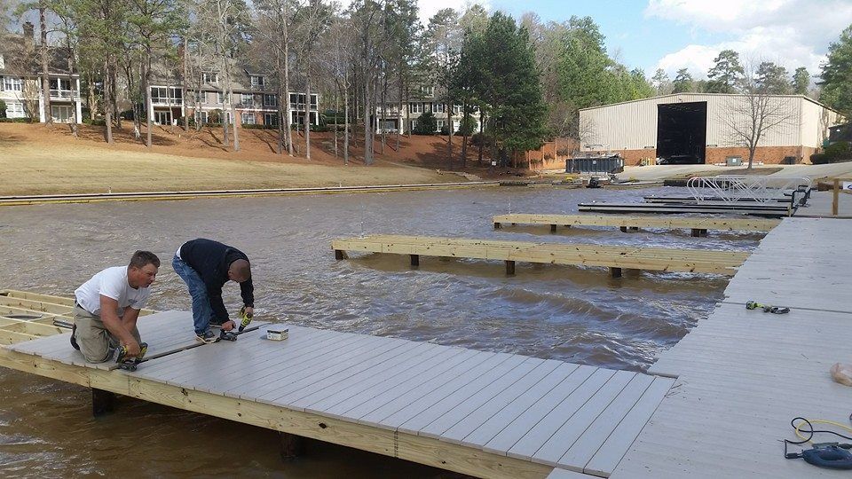 Two people installing a dock on a lake. Brown and gray dock materials. Buildings in the background.