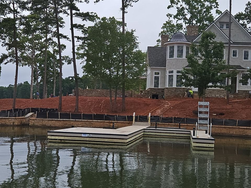 Dock on a lake with a house under construction in the background; dirt, trees and a cloudy sky.