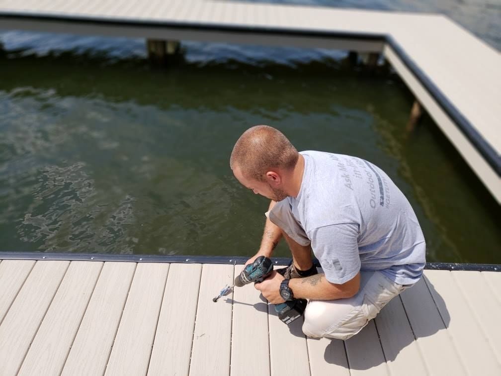 Man kneeling on a dock, using a drill to secure a plank, water in the background.