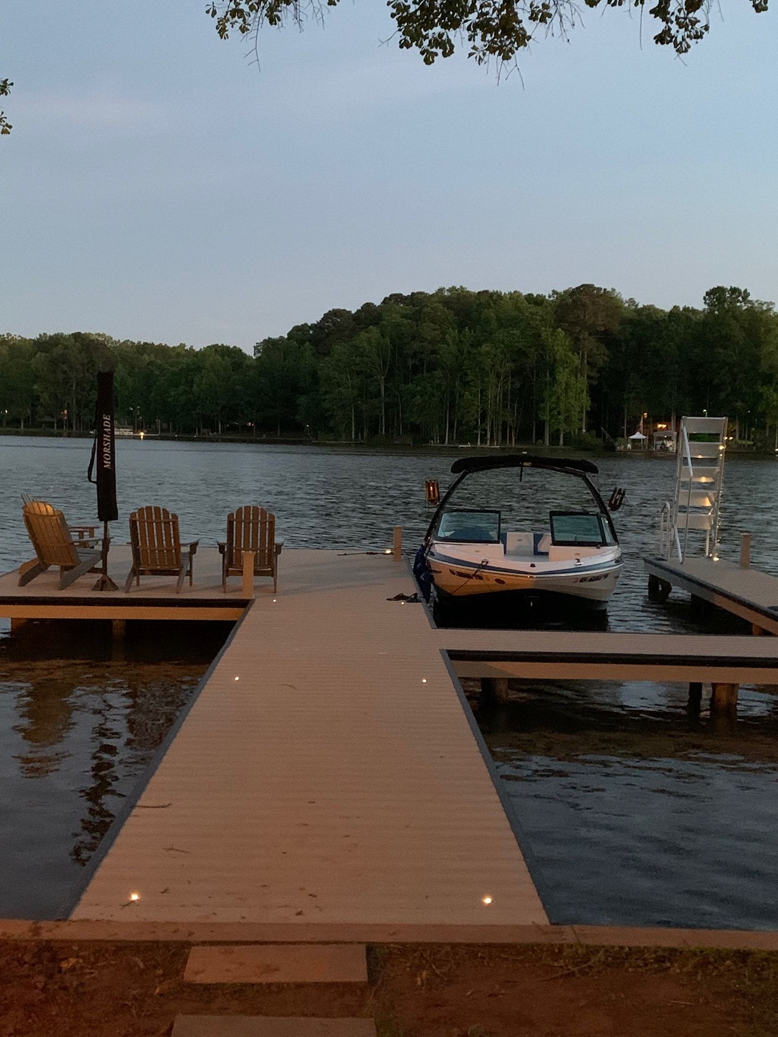 Wooden dock extending into lake, boat docked, chairs, trees in background, overcast sky.