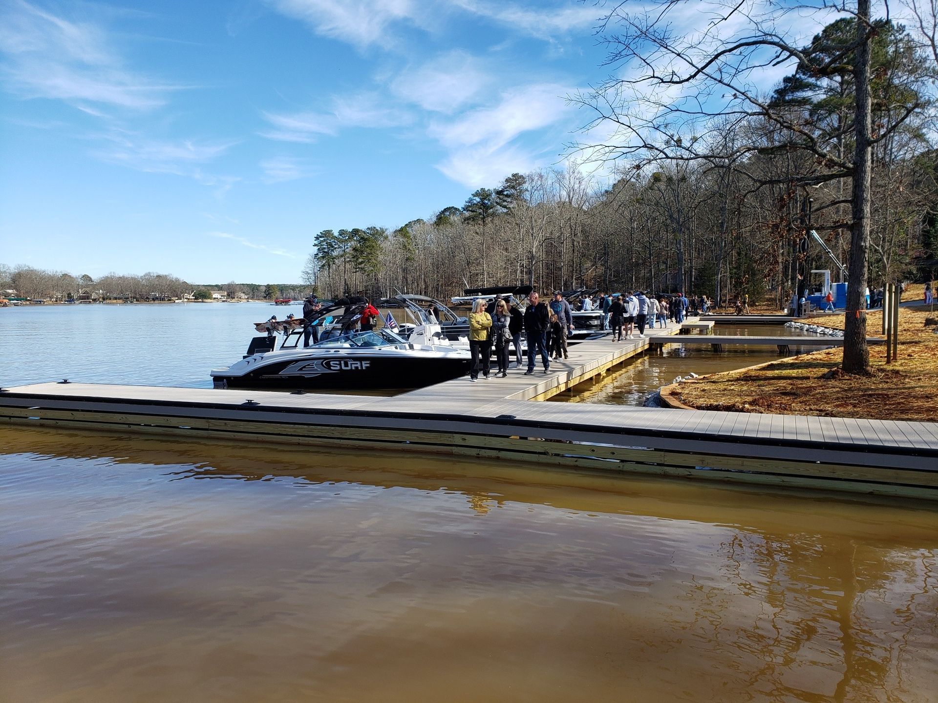 Boat docked at a pier, people walking along it. Trees and lake in the background. Bright sunny day.