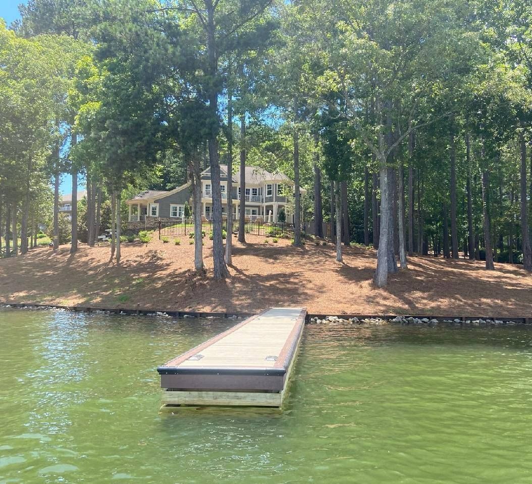 Dock in foreground, lake and trees with a large house visible in the background on a sunny day.