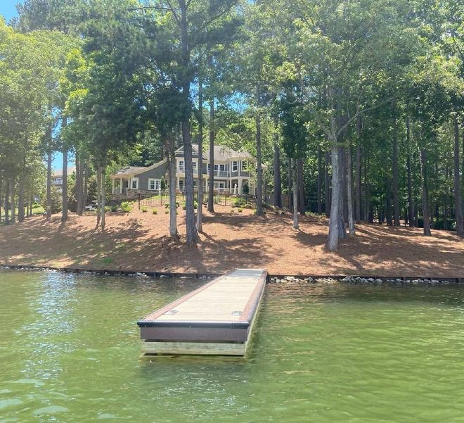 Dock in foreground, lake and trees with a large house visible in the background on a sunny day.
