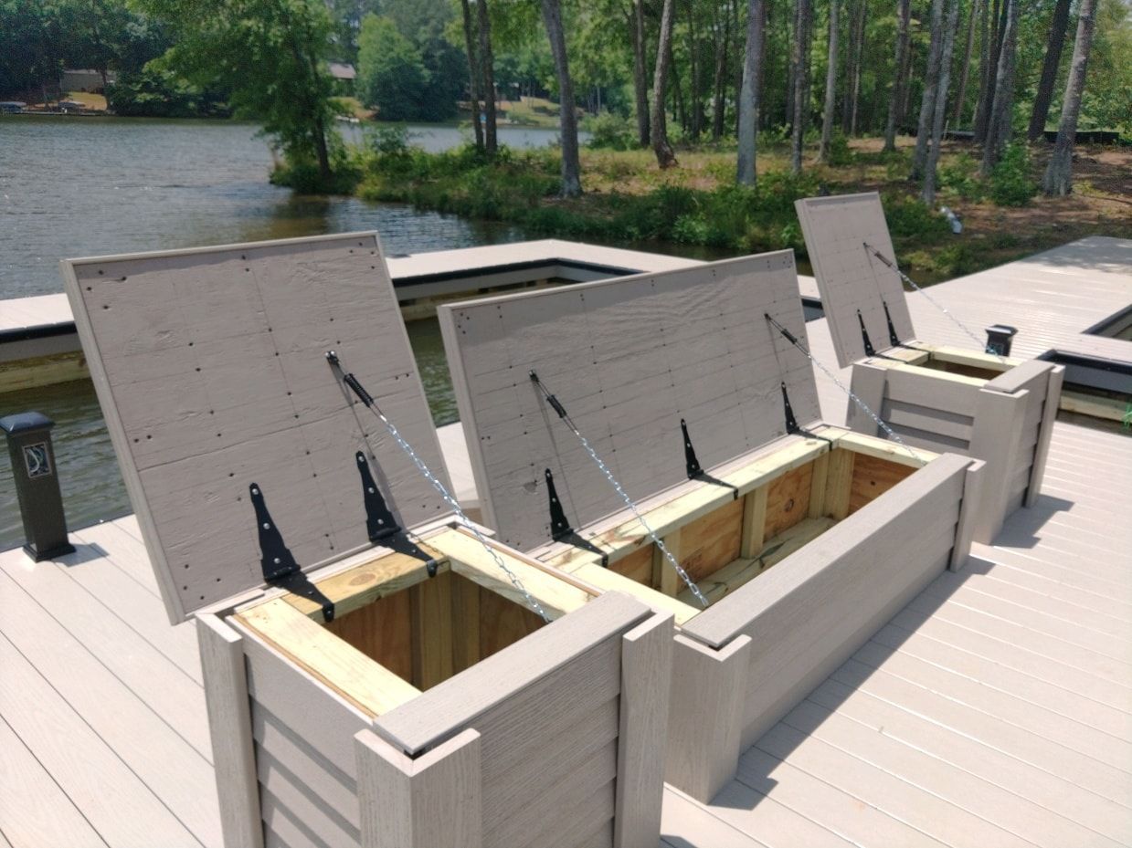 Three gray storage benches with open lids on a wooden dock overlooking water and trees.