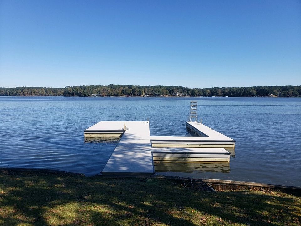 Floating dock on a calm lake with a ladder leading up, green shoreline, and forested background under a blue sky.