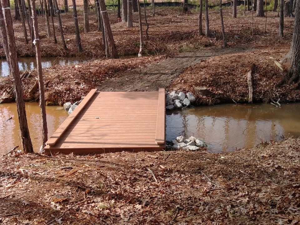 Wooden footbridge over a small creek in a wooded area. Brown leaves and trees surround the bridge.