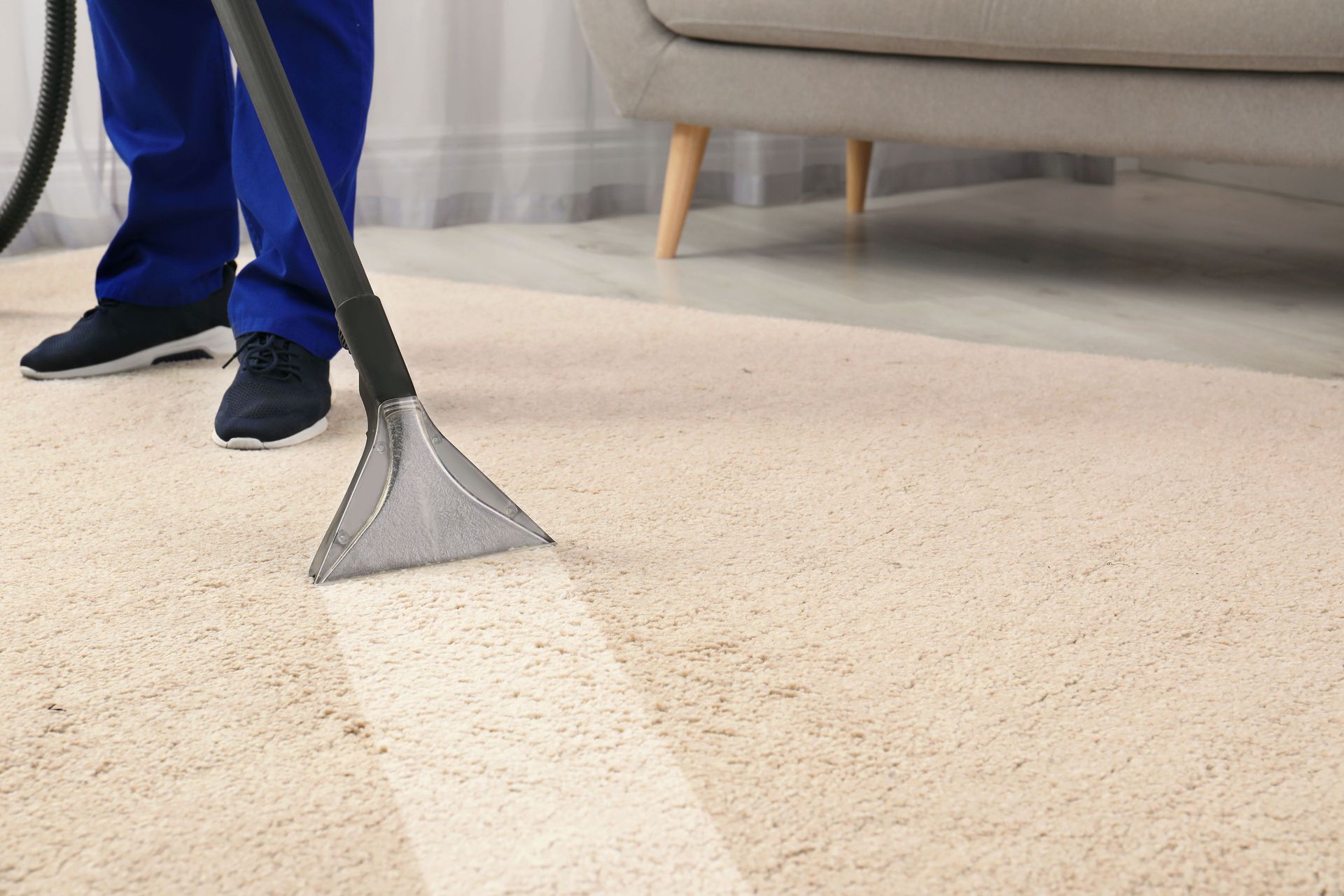 Vacuuming a beige carpet beside a couch, with a blue-clad person visible from the knees down