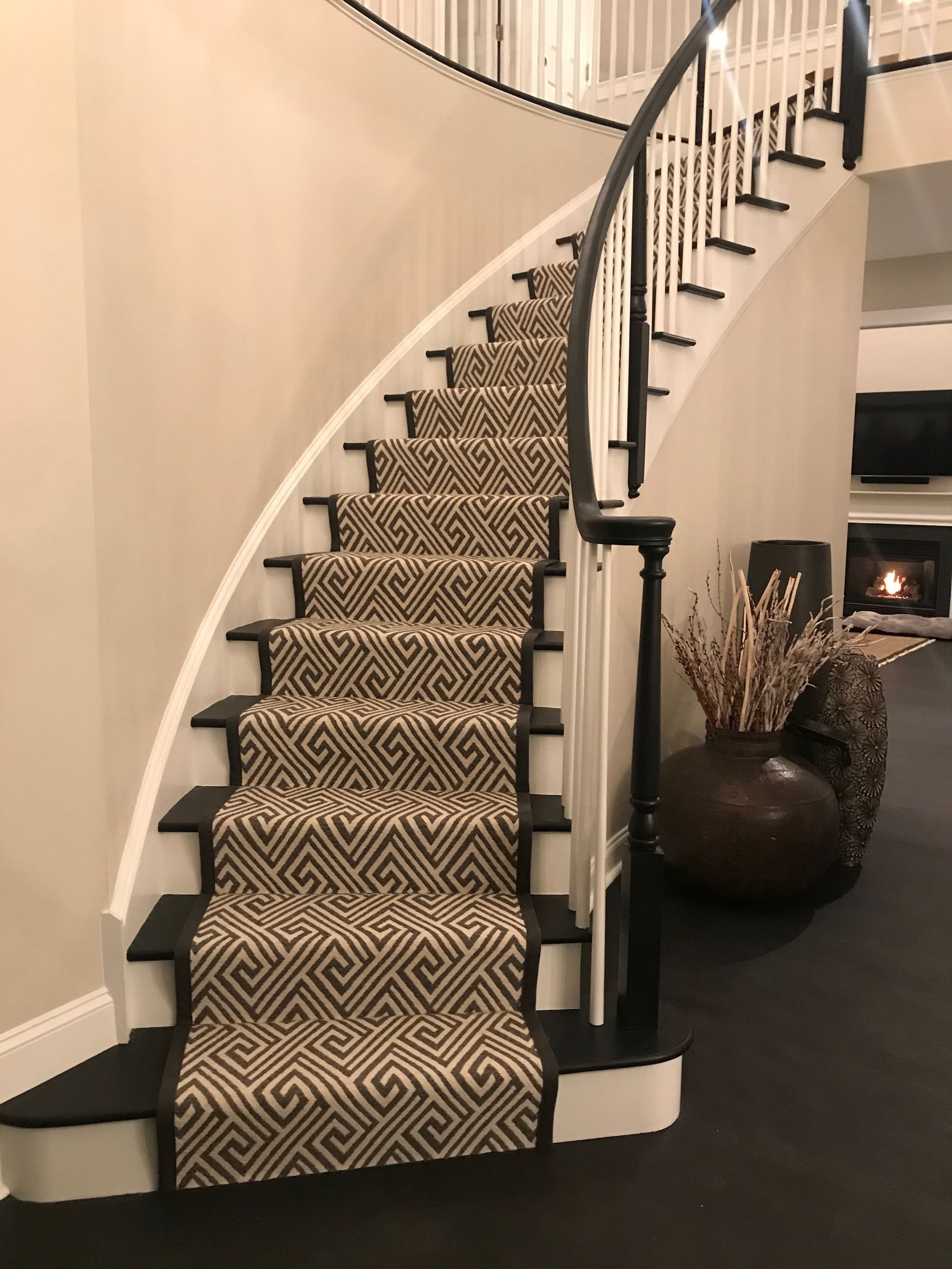 Curved carpeted staircase with black railing in a beige foyer, beside a large decorative vase.