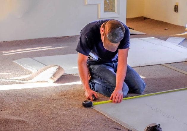 Person kneeling on carpet, measuring and cutting flooring with a tape measure and utility knife.