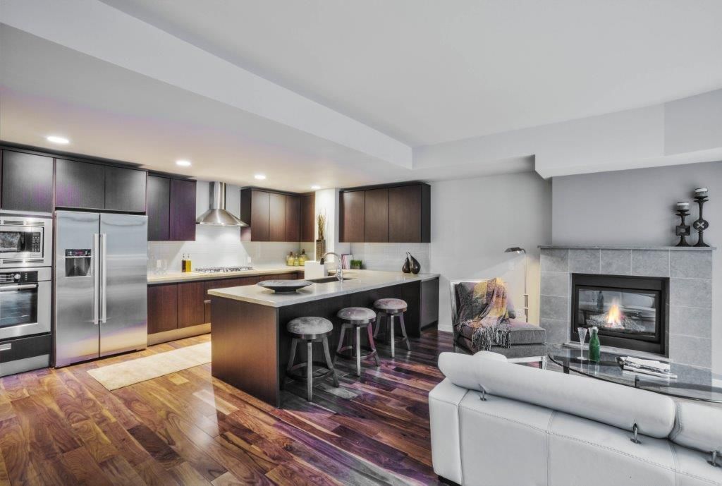 Modern open-concept kitchen and living room with dark wood cabinets, island stools, fireplace, and white sofa.