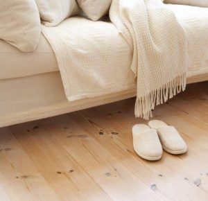 Cream slippers beside a bed with a beige blanket on a wooden floor.