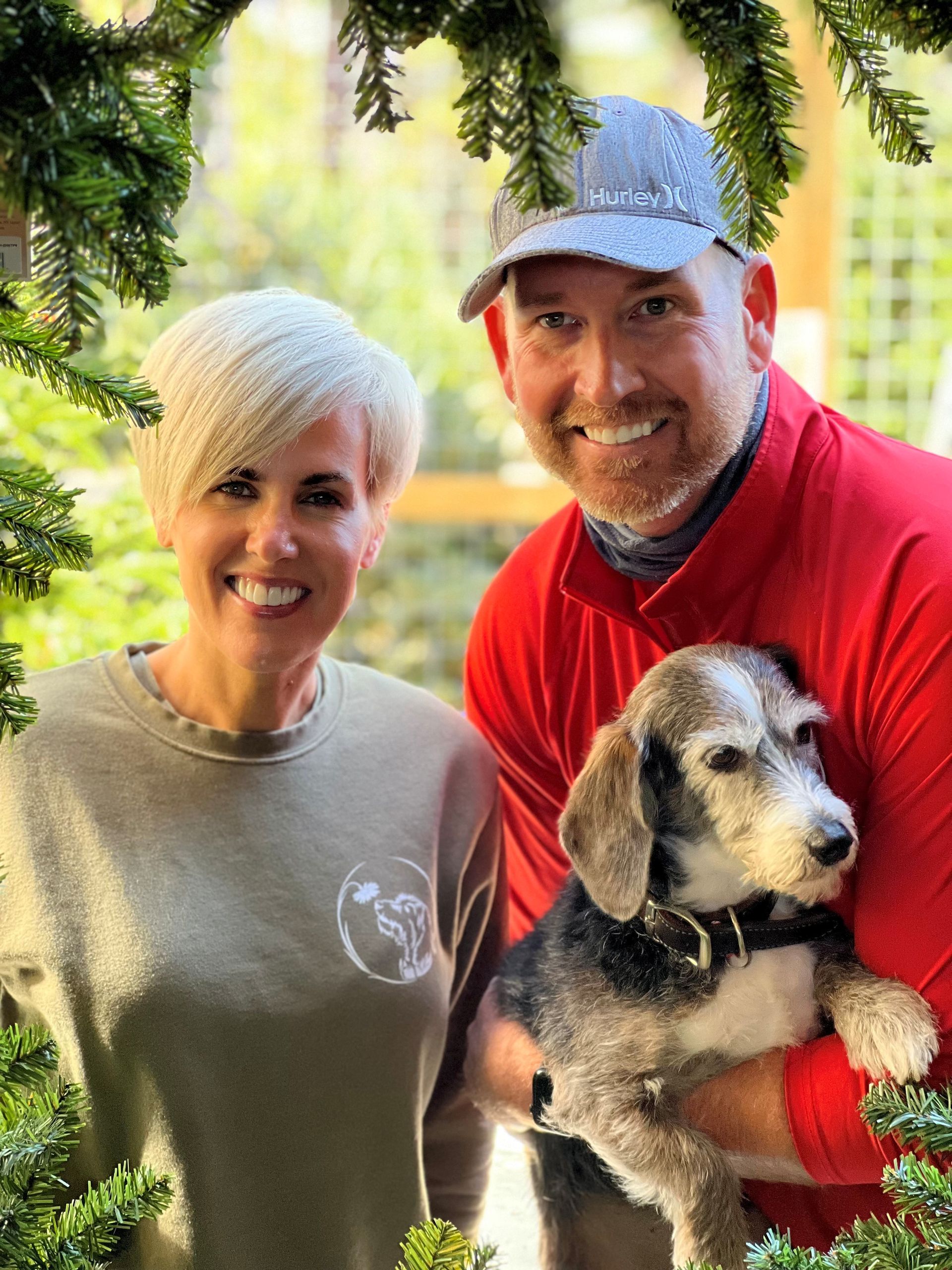 Couple smiling, holding a dog framed by greenery; man in red, woman in green.