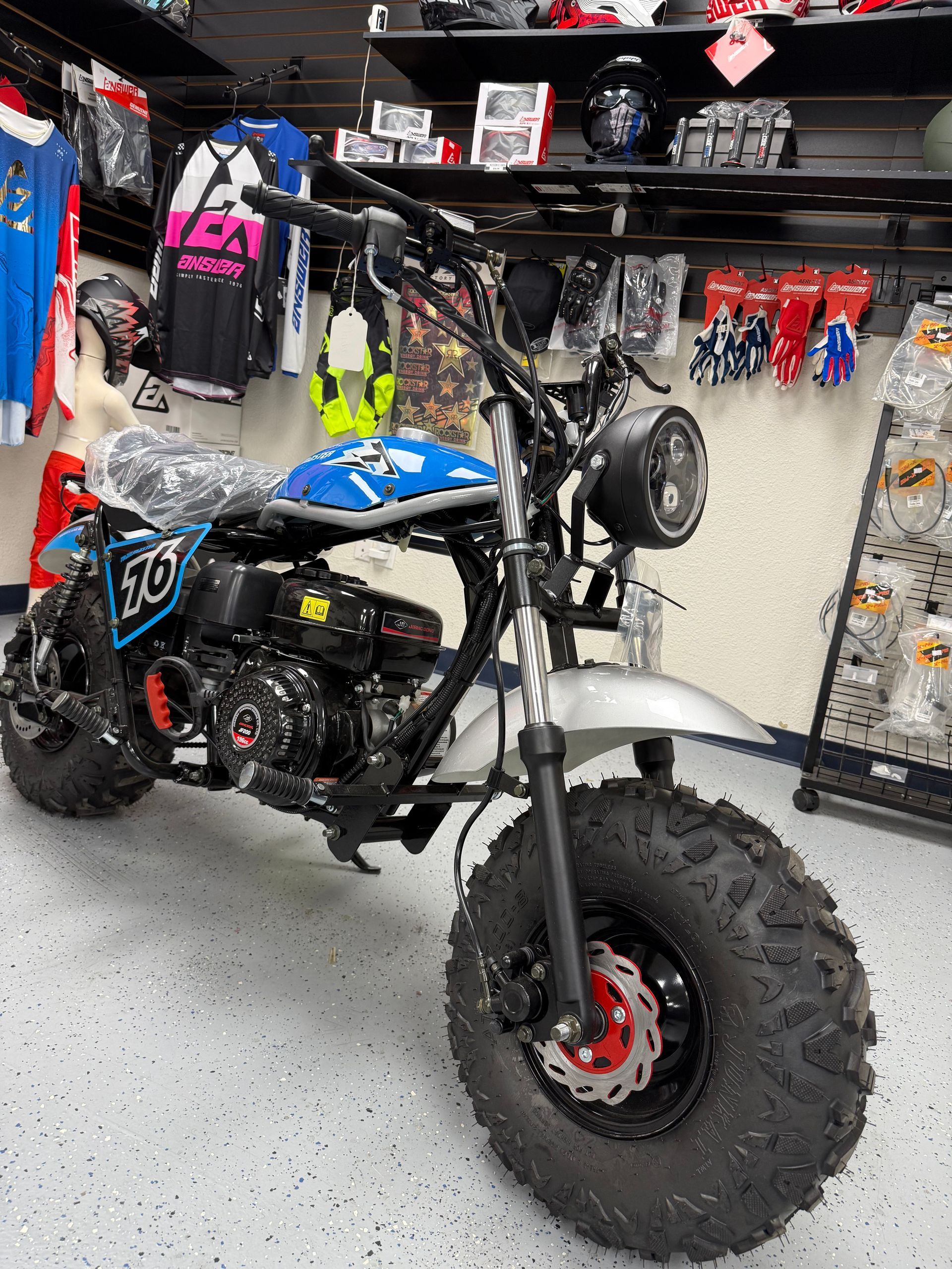 Black-and-blue mini dirt bike with knobby tires in a garage, surrounded by gear and hanging riding clothes