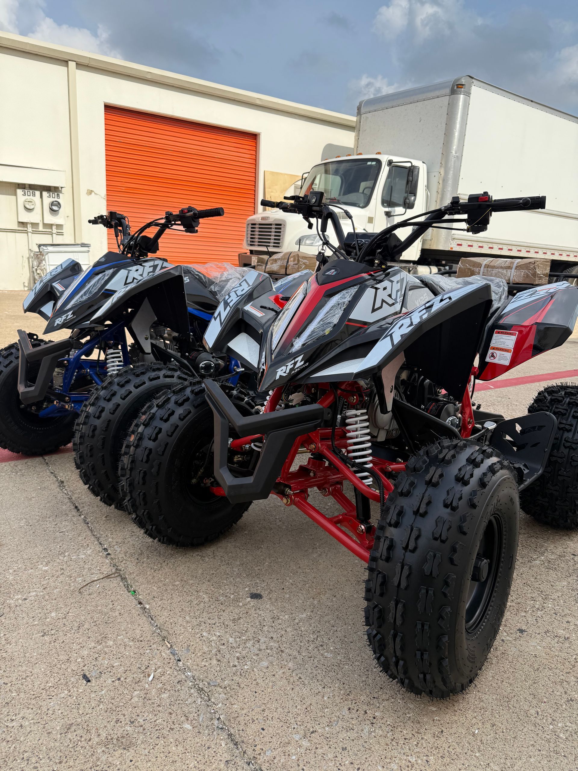 Two sport ATVs parked outside a building, one black and one white-red, on a concrete lot.