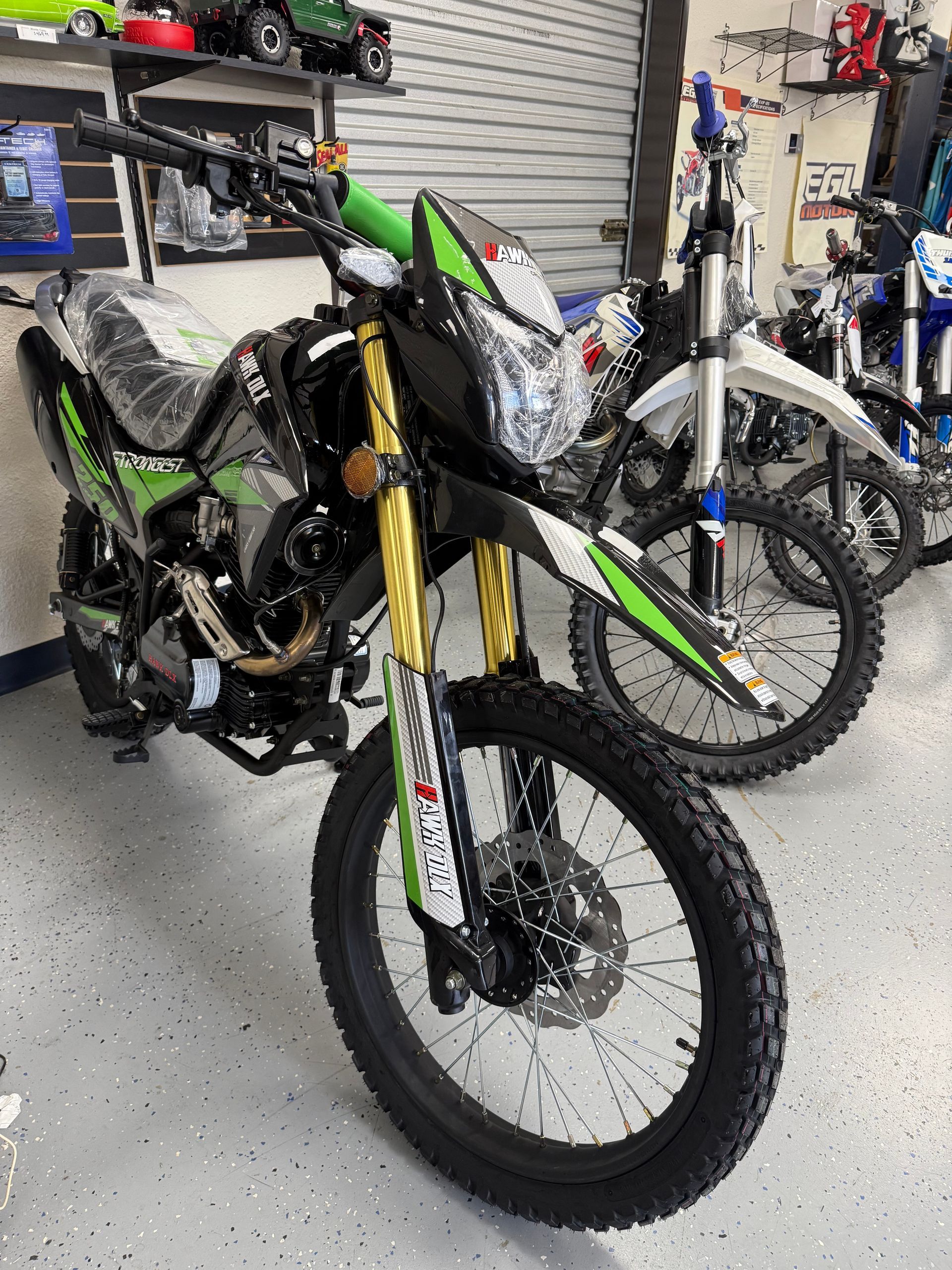 Green and black off-road motorcycle displayed indoors in a bike shop, with other bikes behind it