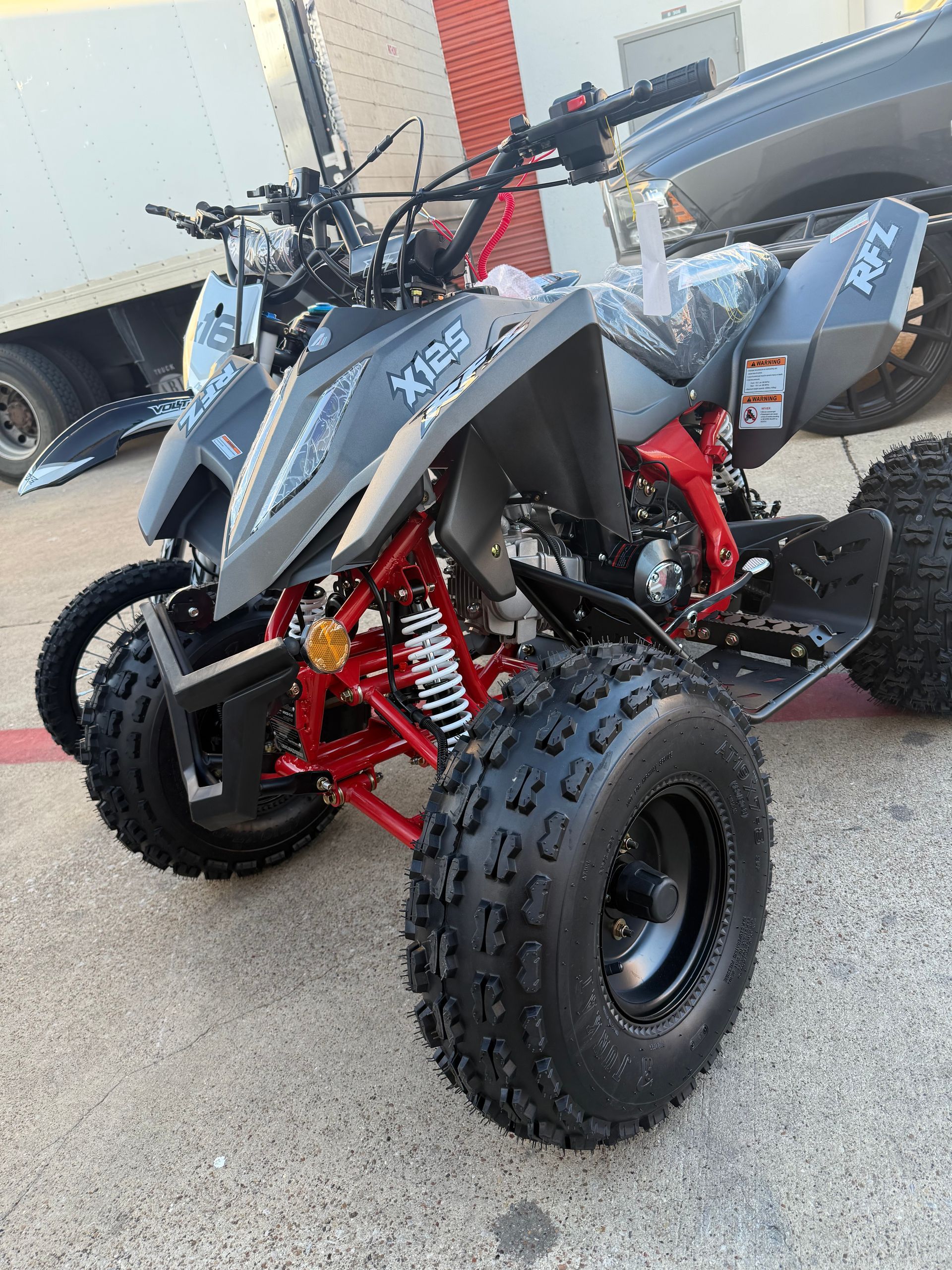 Gray and red off-road ATV parked on pavement with knobby tires and a black seat, near a trailer