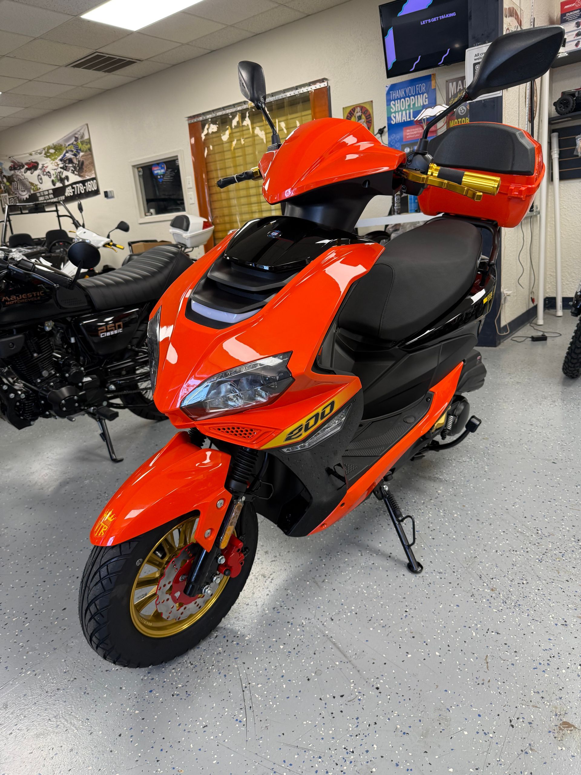 Orange scooter on display in a showroom, with a black seat and gold wheels.