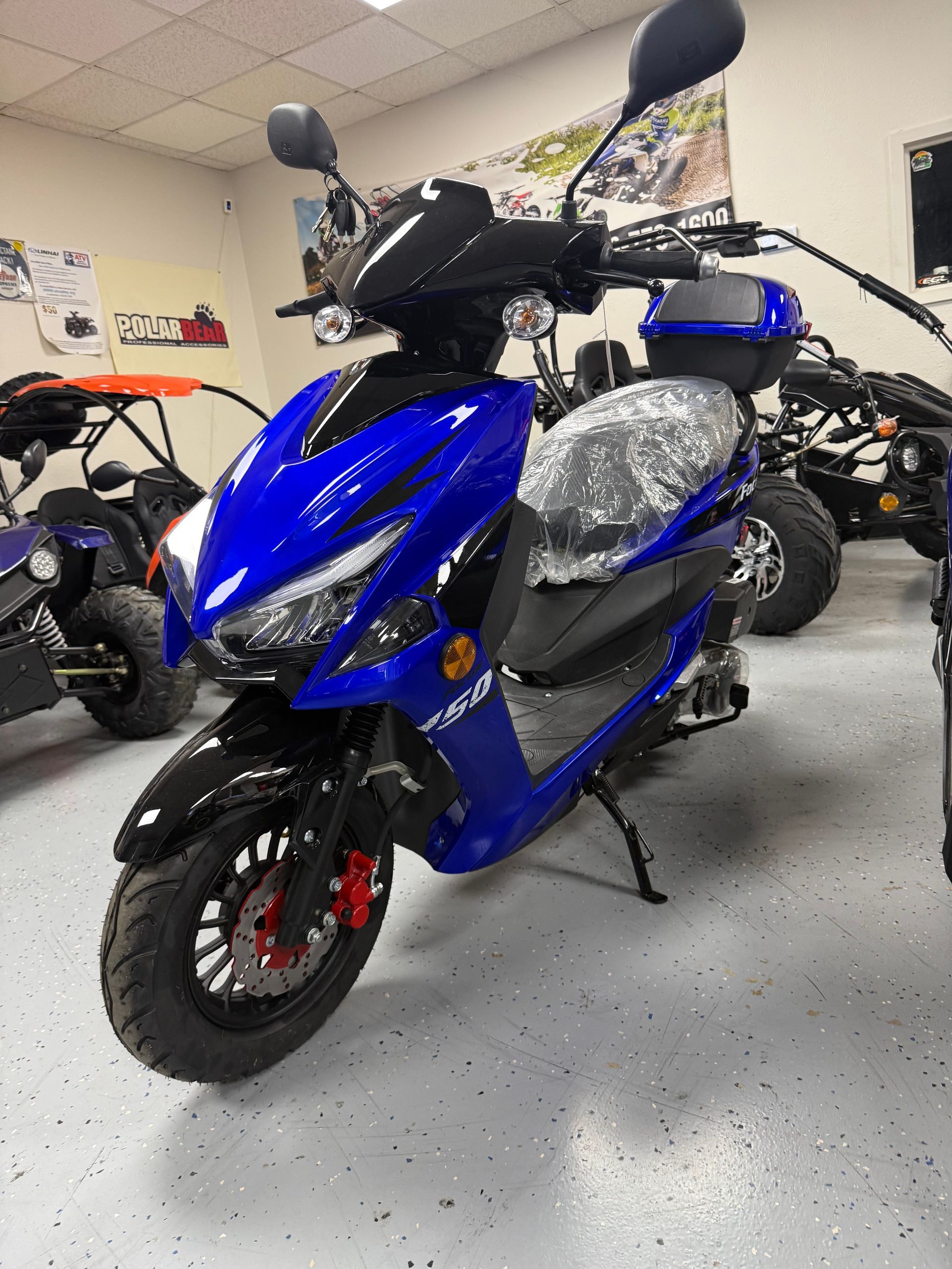 Blue scooter in a showroom, wrapped in protective plastic, with other motorcycles in the background.