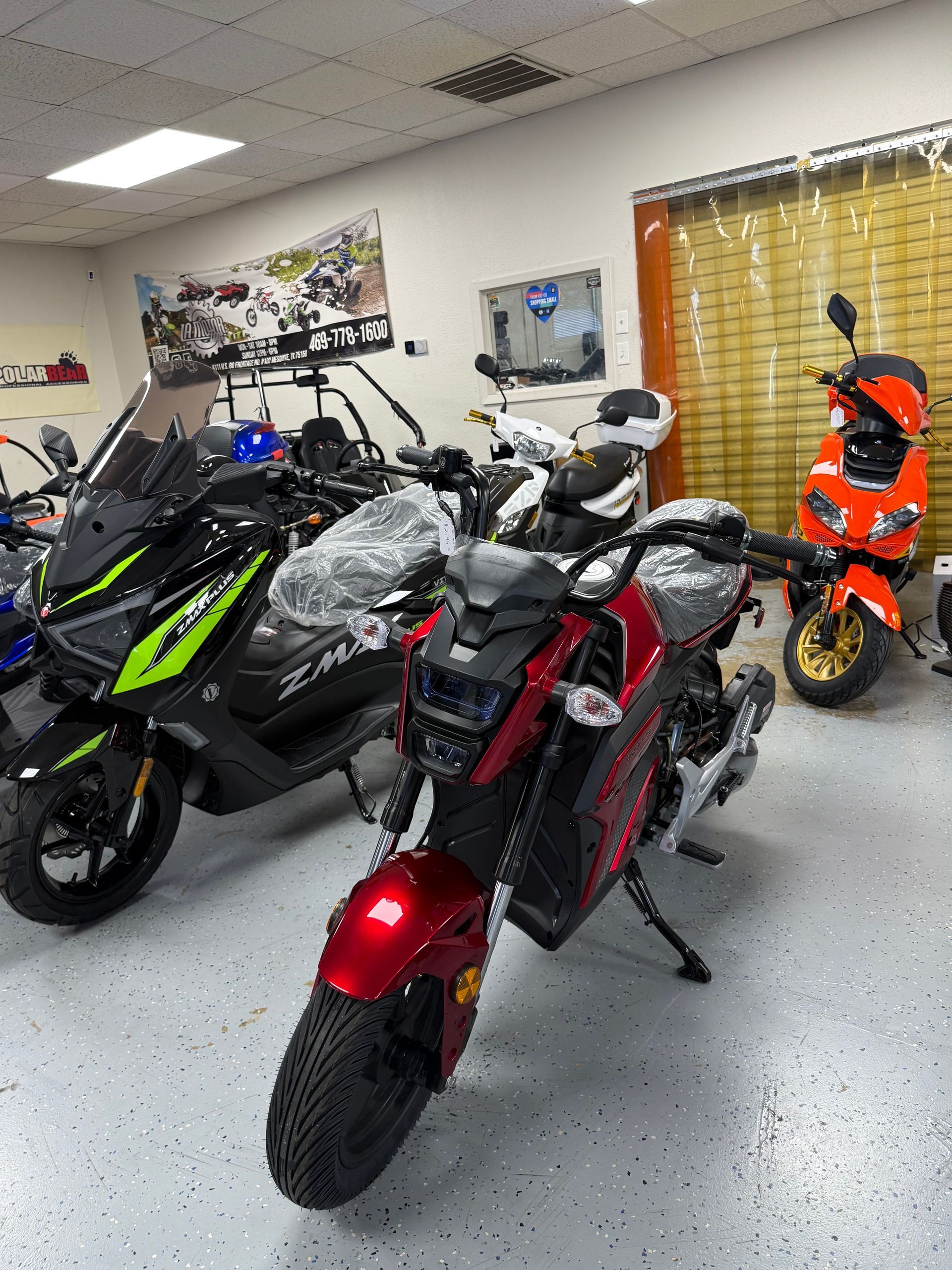 Three motorcycles in a showroom, including a red bike in front and an orange mini bike by the window.