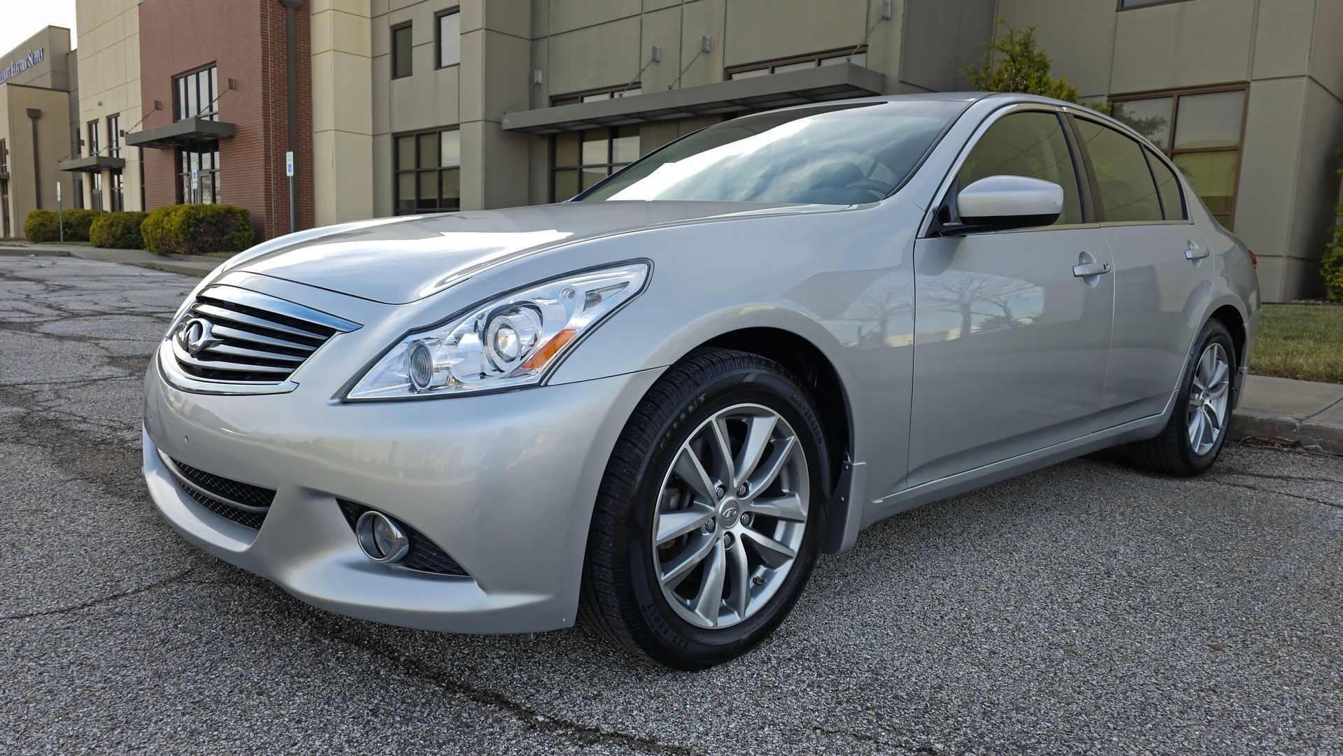 Silver Infiniti G37 sedan parked on gravel in front of a modern building.