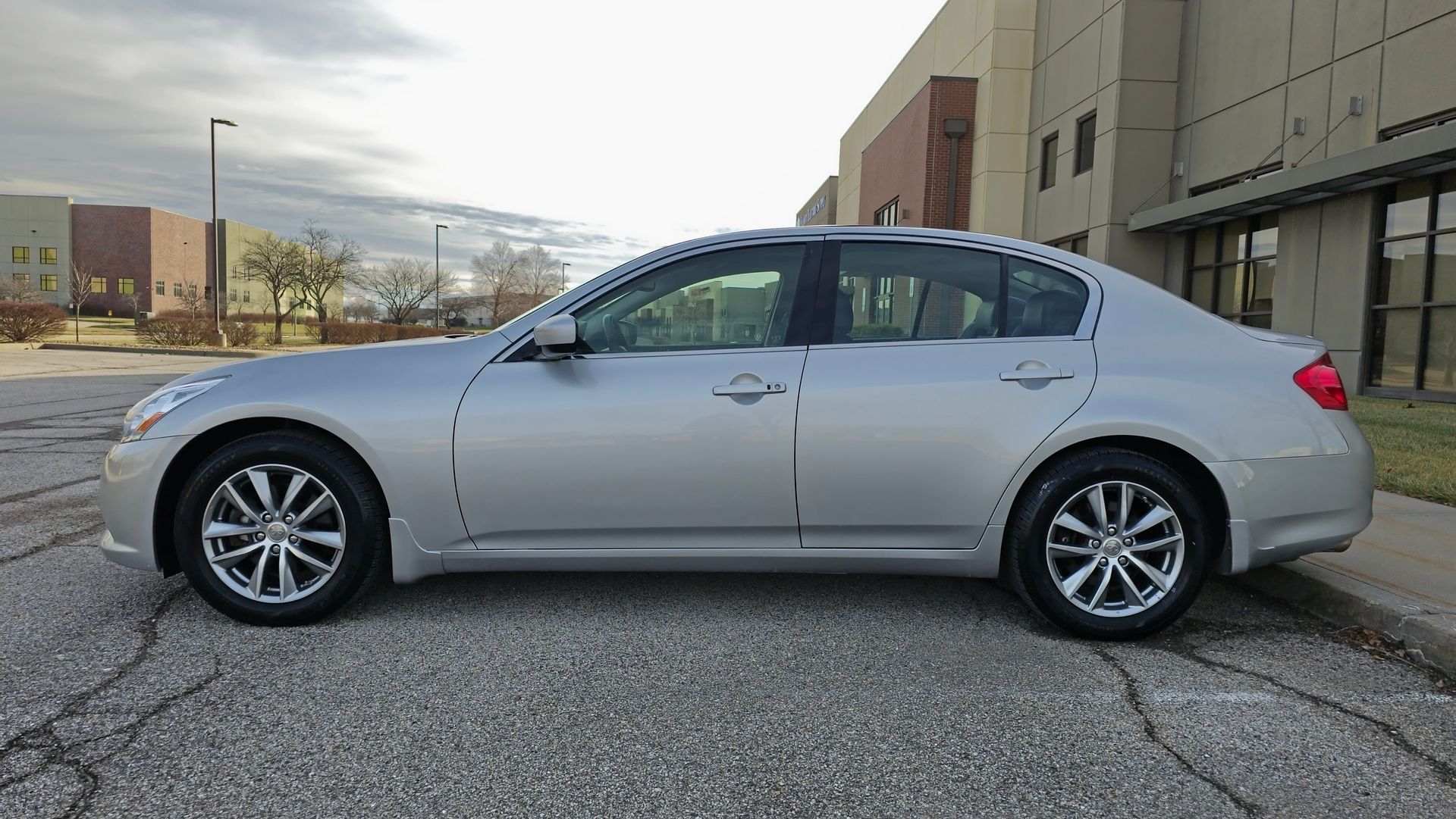 Silver Infiniti G37 sedan parked on cracked asphalt in front of commercial buildings.